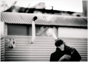 Black and white street portrait of a man exhaling smoke in front of a trailer park with corrugated metal siding