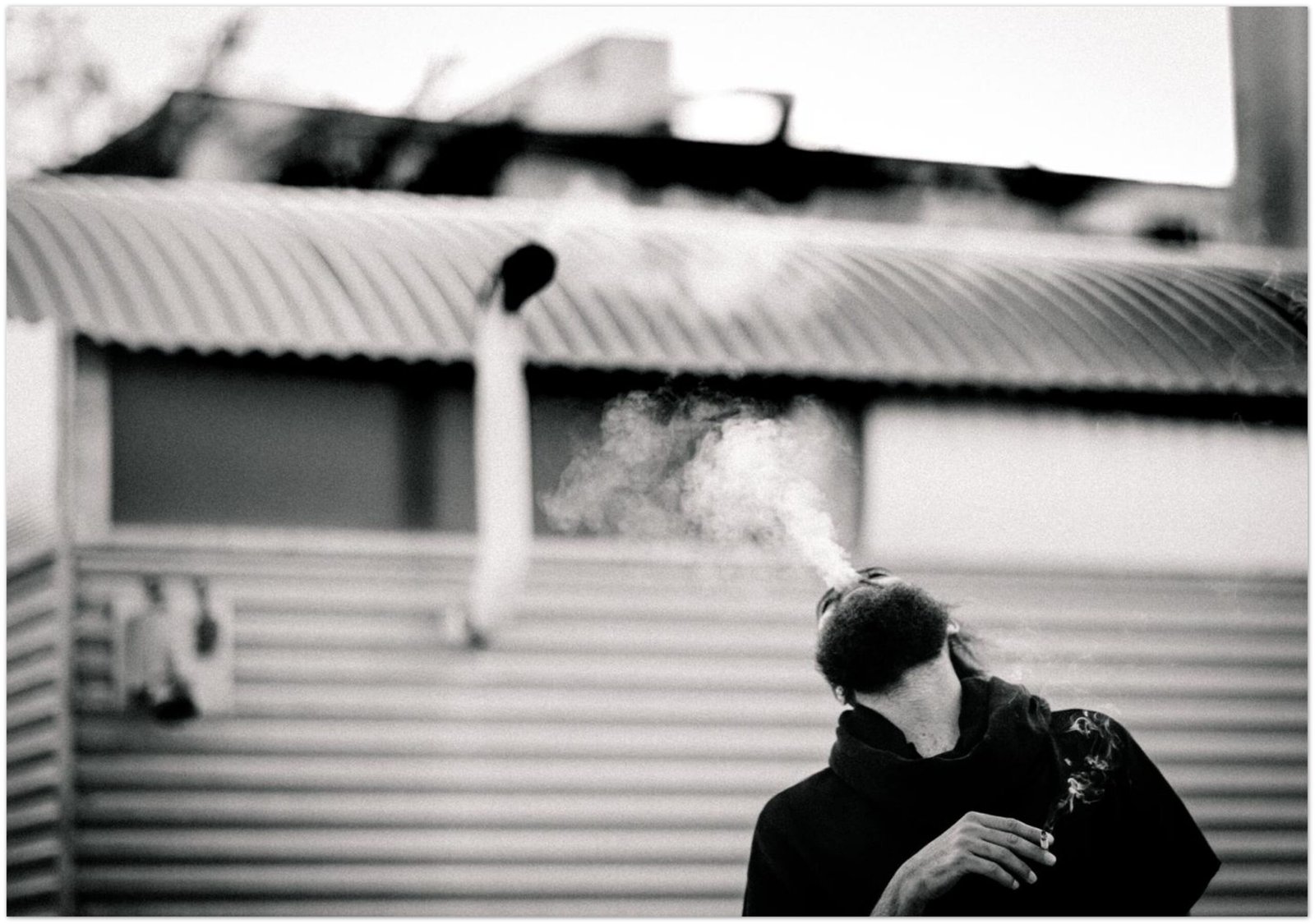 Black and white street portrait of a man exhaling smoke in front of a trailer park with corrugated metal siding