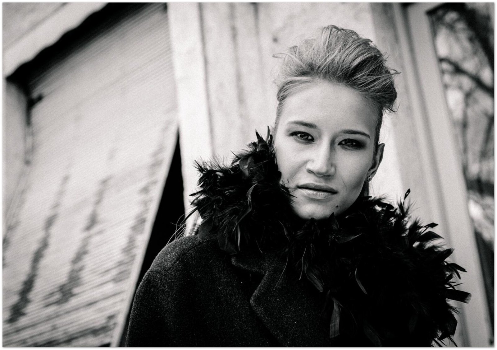 Simple Black and white fashion portrait of a woman with feather collar and windswept hair against weathered wooden Scandinavian facade