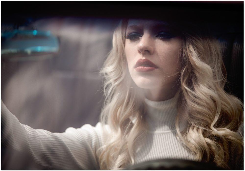 Cinematic portrait of a blonde woman with wavy hair behind the wheel of a vintage car, soft natural light, dreamy color tones